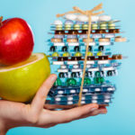 Human hand holding stack of pills and fruits on blue.