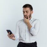 confused handsome man puts hand on chin holding and looking at phone isolated on white background with copy space
