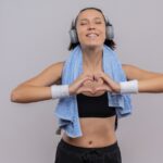 young fitness girl in sportswear with headphones on head and towel on her neck making heart gesture with fingers with closed eyes feeling positive emotions standing over white background