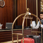 Young bellboy in uniform pushing cart with suitcases along reception counter