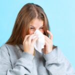 Portrait of young girl sneezing isolated over blue background. Cold and flu. Winter sickness