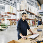 Medium wide shot of male warehouse worker checking orders at computer workstation in warehouse
