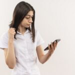 young girl in white shirt looking at screen of her mobile looking confused standing over white background