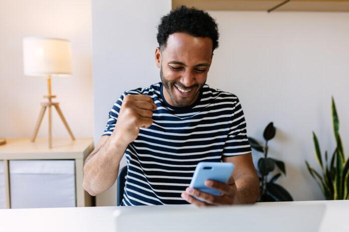 Excited young african american man winning on internet lottery betting online on smartphone app. Millennial guy celebrating victory enjoying success sports bet on cellphone.